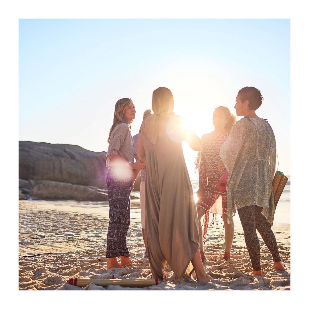A group of women stand together on a sandy beach near the ocean on a bright day, possibly after yoga or a similar activity.