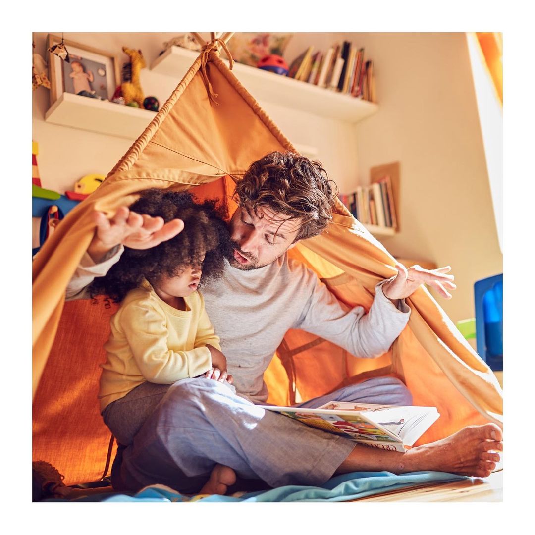 Father and child reading a book inside a teepee in a brightly lit room, creating a heartwarming, playful indoor scene.