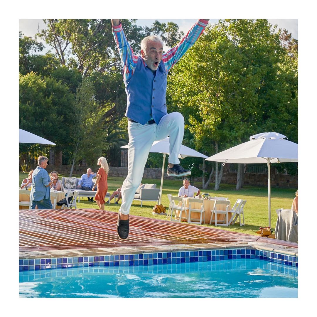 Enthusiastic senior man mid-air, jumping joyfully near a pool at an outdoor party, surrounded by casually dressed guests and lush greenery.