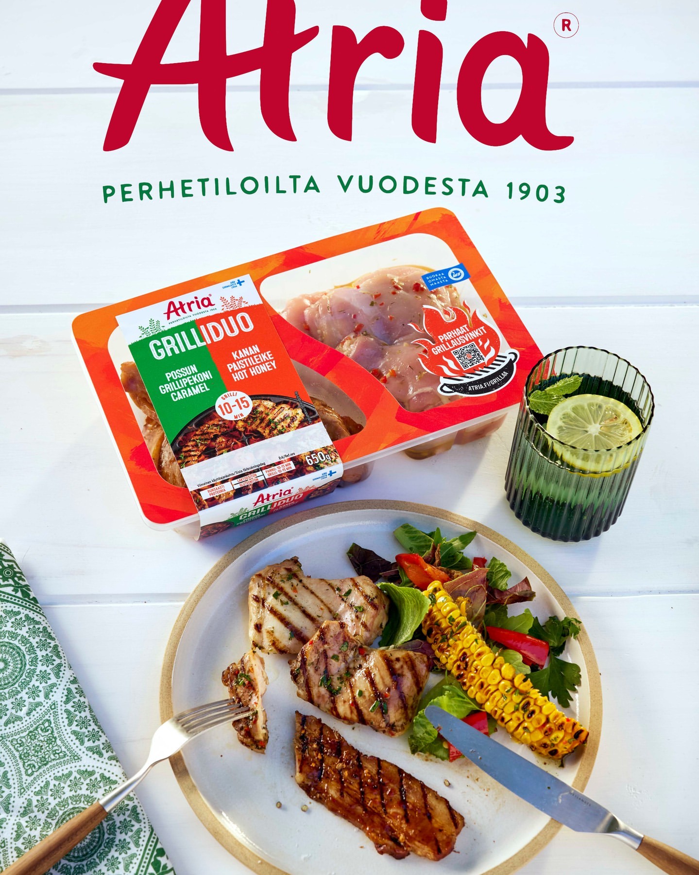 Prepared Atria grilled chicken meal with corn, salad, and a glass of lemonade, presented on a white wooden table with a green napkin.