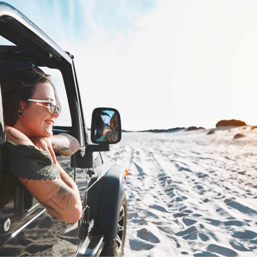 A smiling woman with tattoos and sunglasses leans out of an off-road vehicle window, enjoying a sunny day at the beach with sand dunes.