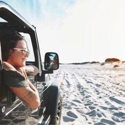 Smiling woman wearing sunglasses leaning out of a vehicle window in a sandy desert landscape.