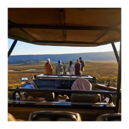 Group of six people standing behind an open safari vehicle looking out across a wide African landscape at sunset.