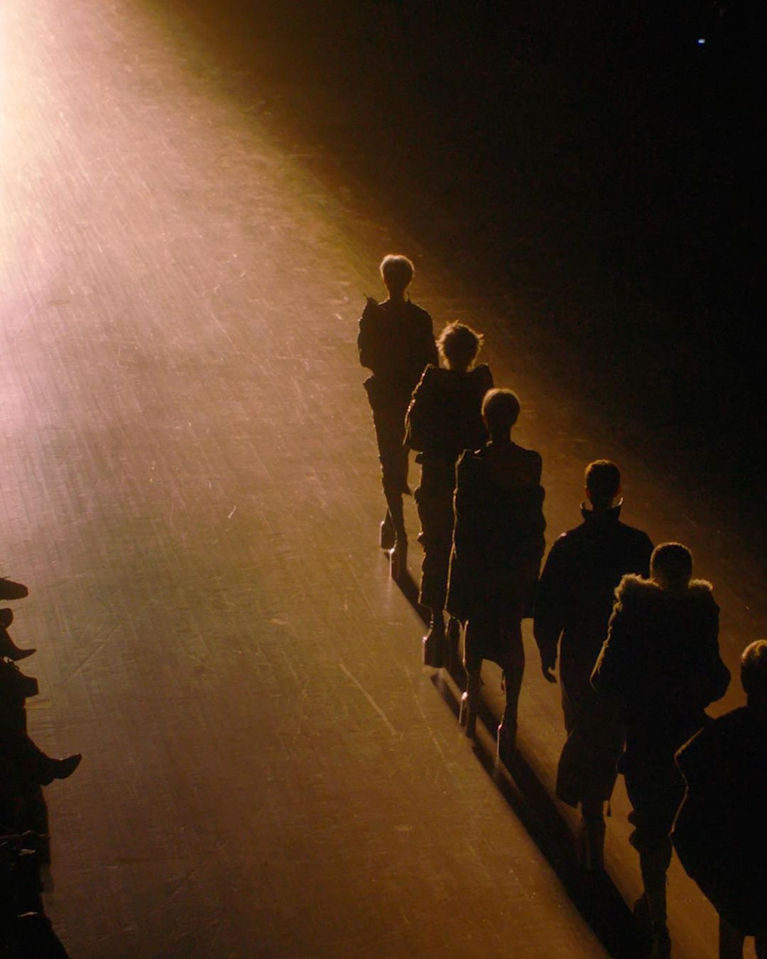 Fashion show runway with silhouettes of models walking in single file under a spotlight. Dark, warm-toned aesthetic.