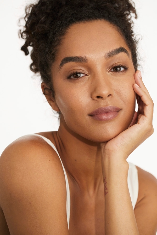 Portrait of a beautiful young woman with curly hair resting her chin on her hand, looking directly at the camera against a white background.
