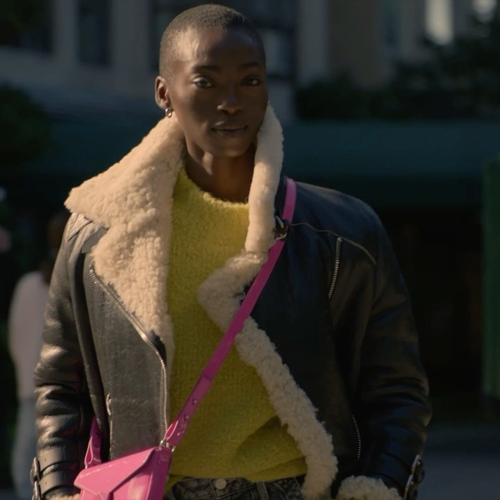 Stylish woman with buzzcut wears yellow sweater, shearling jacket, and pink purse in an urban setting with neutral background tones.