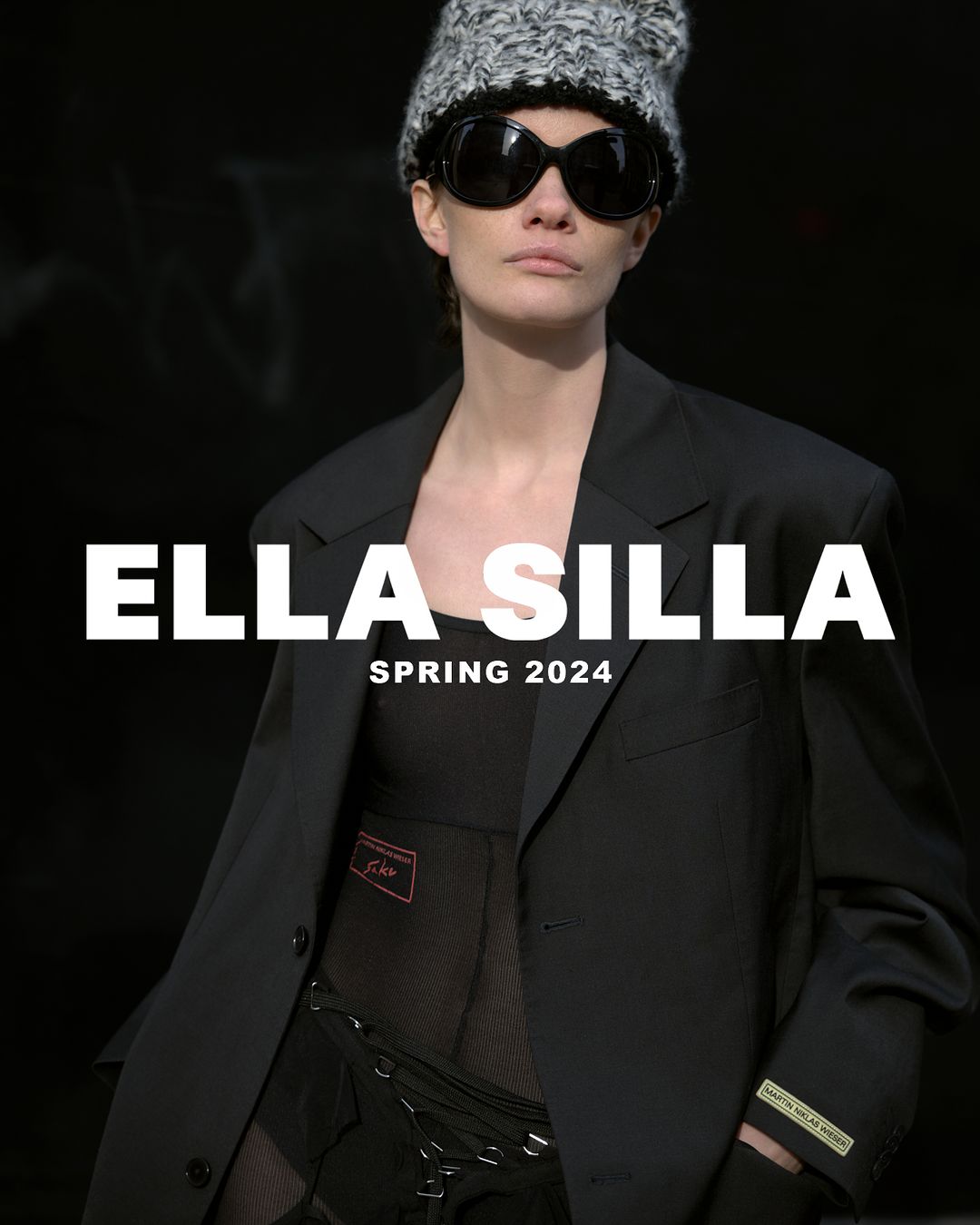 Fashion shot of a woman wearing a black blazer, knit hat, and sunglasses against a dark background with the text 'Ella Silla Spring 2024'.