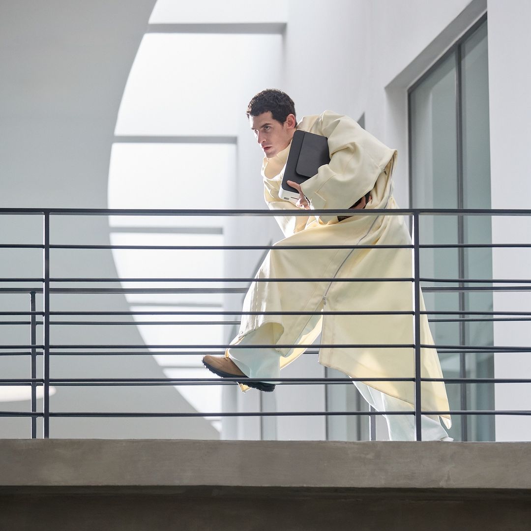 Man in robe runs on balcony indoors holding folder with sunlight and architectural elements providing a dynamic composition.