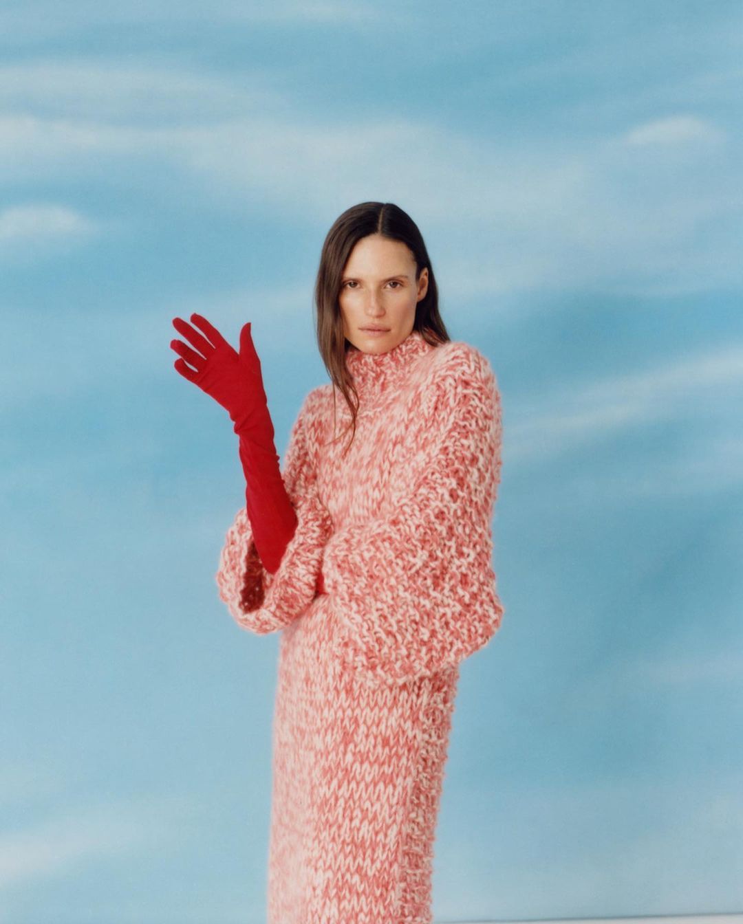 Woman in a pink knit dress and red gloves poses in front of a light blue background with wispy clouds, fashion photography, editorial.
