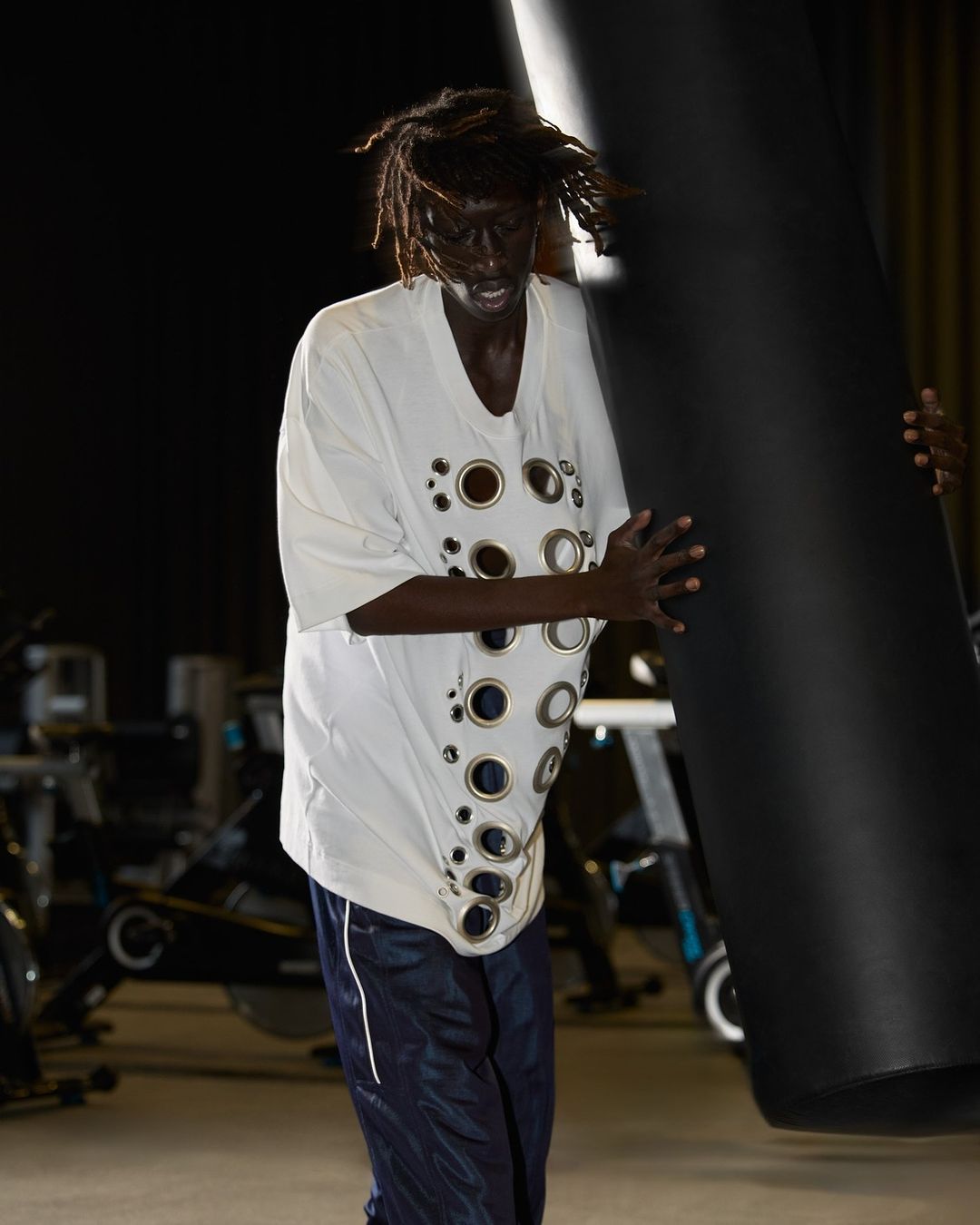 An adult male with dreadlocks is seen in a fashion-forward t-shirt and athletic pants, indoors at a gym with exercise bikes.