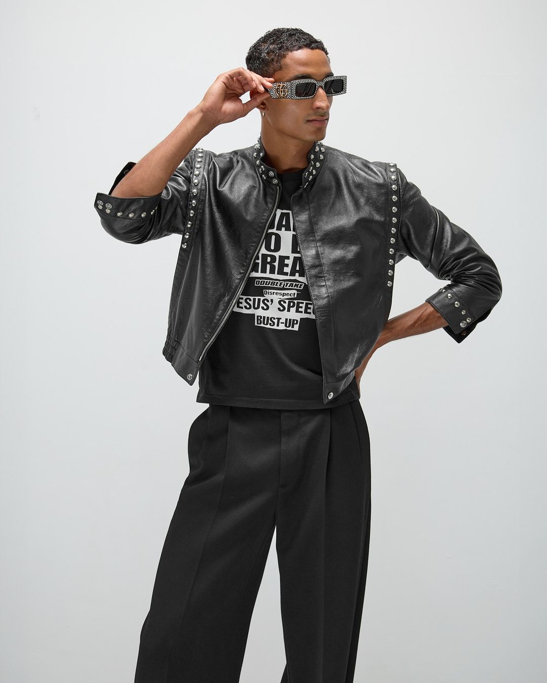 Adult male in fashionable black leather jacket, sunglasses, and wide-leg pants striking a confident pose in a studio setting.