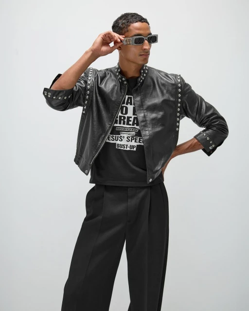 Adult male in fashionable black leather jacket, sunglasses, and wide-leg pants striking a confident pose in a studio setting.