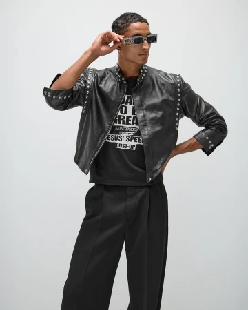 Adult male in fashionable black leather jacket, sunglasses, and wide-leg pants striking a confident pose in a studio setting.