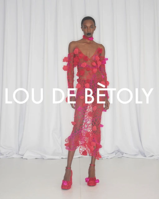 A slender Black woman in a sheer pink floral gown poses against a white curtain backdrop, showcasing high-heeled shoes with floral accents.