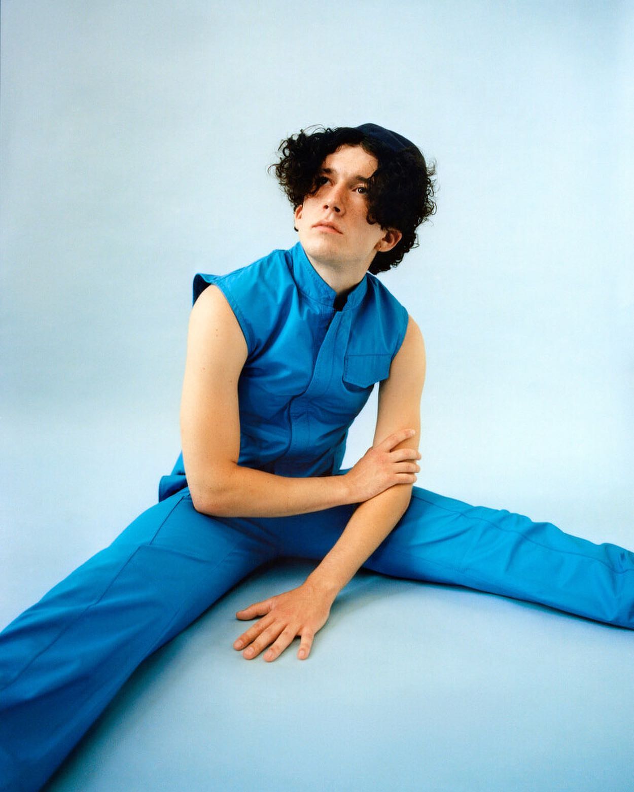 Young adult man in blue sleeveless jumpsuit poses on light blue backdrop, looking up and away with arms crossed, hat on head, indoor.