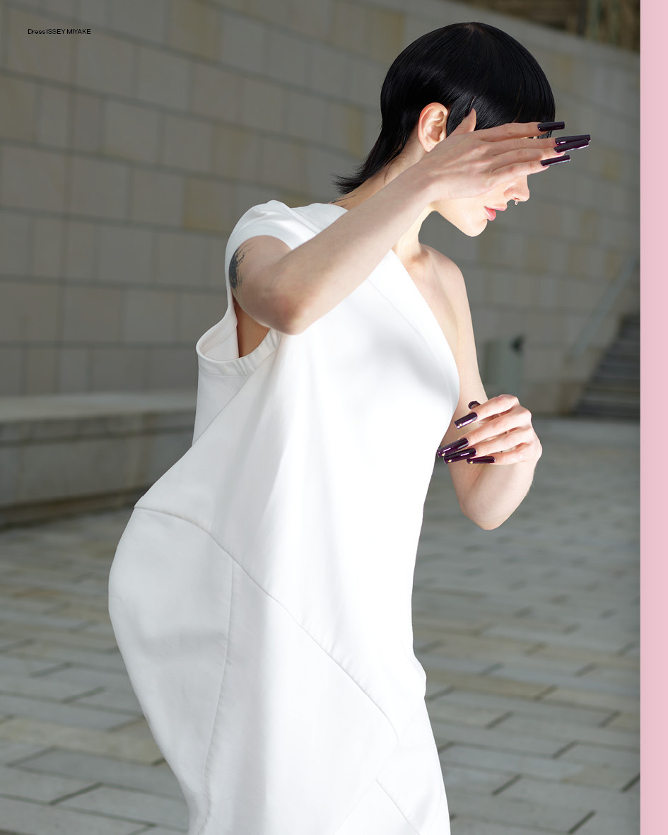 Model with dark hair and long dark fingernails wearing a white dress against a paved outdoor backdrop in Paris.