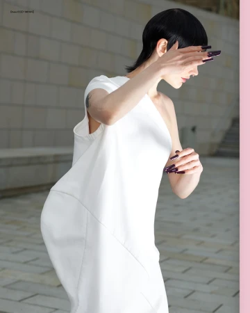 Model with dark hair and long dark fingernails wearing a white dress against a paved outdoor backdrop in Paris.
