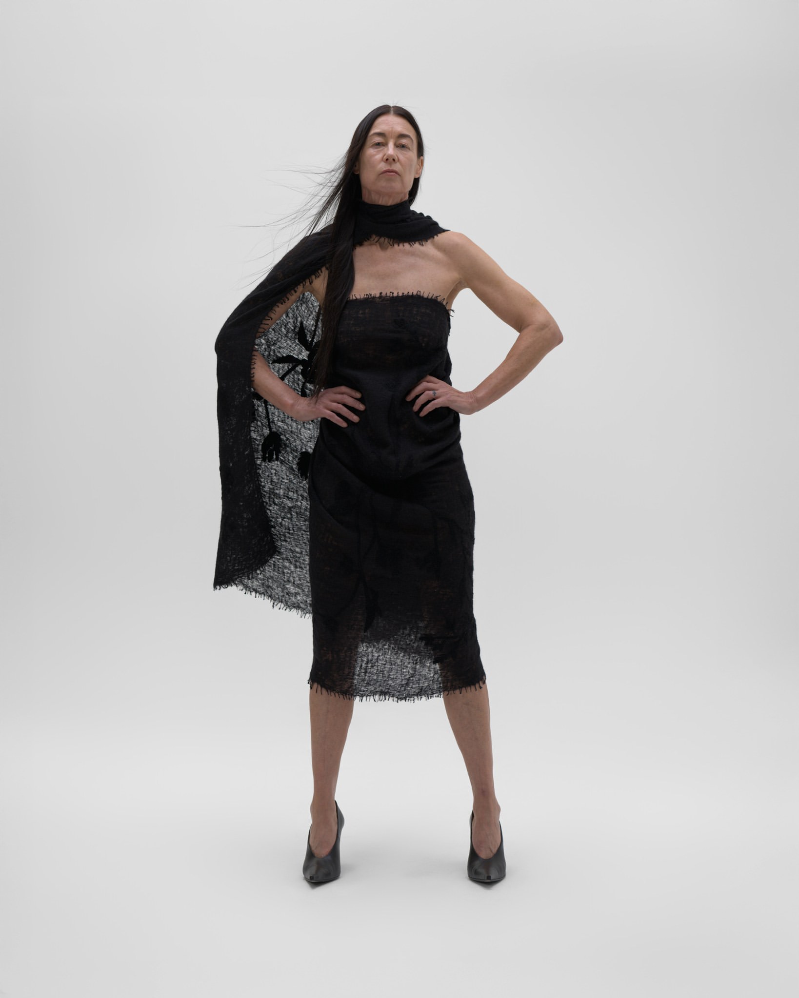Adult female model poses in a black strapless dress with a black lace shawl in a full shot against a seamless white backdrop.