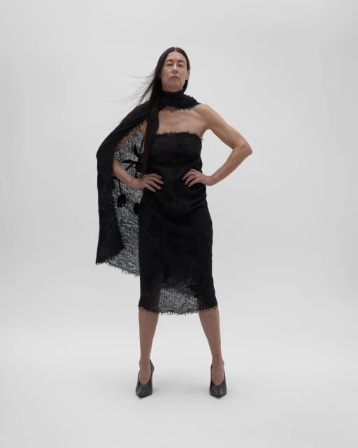Adult female model poses in a black strapless dress with a black lace shawl in a full shot against a seamless white backdrop.