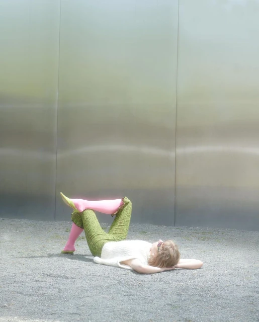 Woman lying on ground in green pants and pink boots against a metal wall.  Blonde hair, white top. Fashion shot in outdoor daylight.