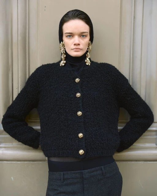 Woman in a black cardigan with decorative gold buttons poses against a neutral door. She wears large gold earrings and a black head covering.