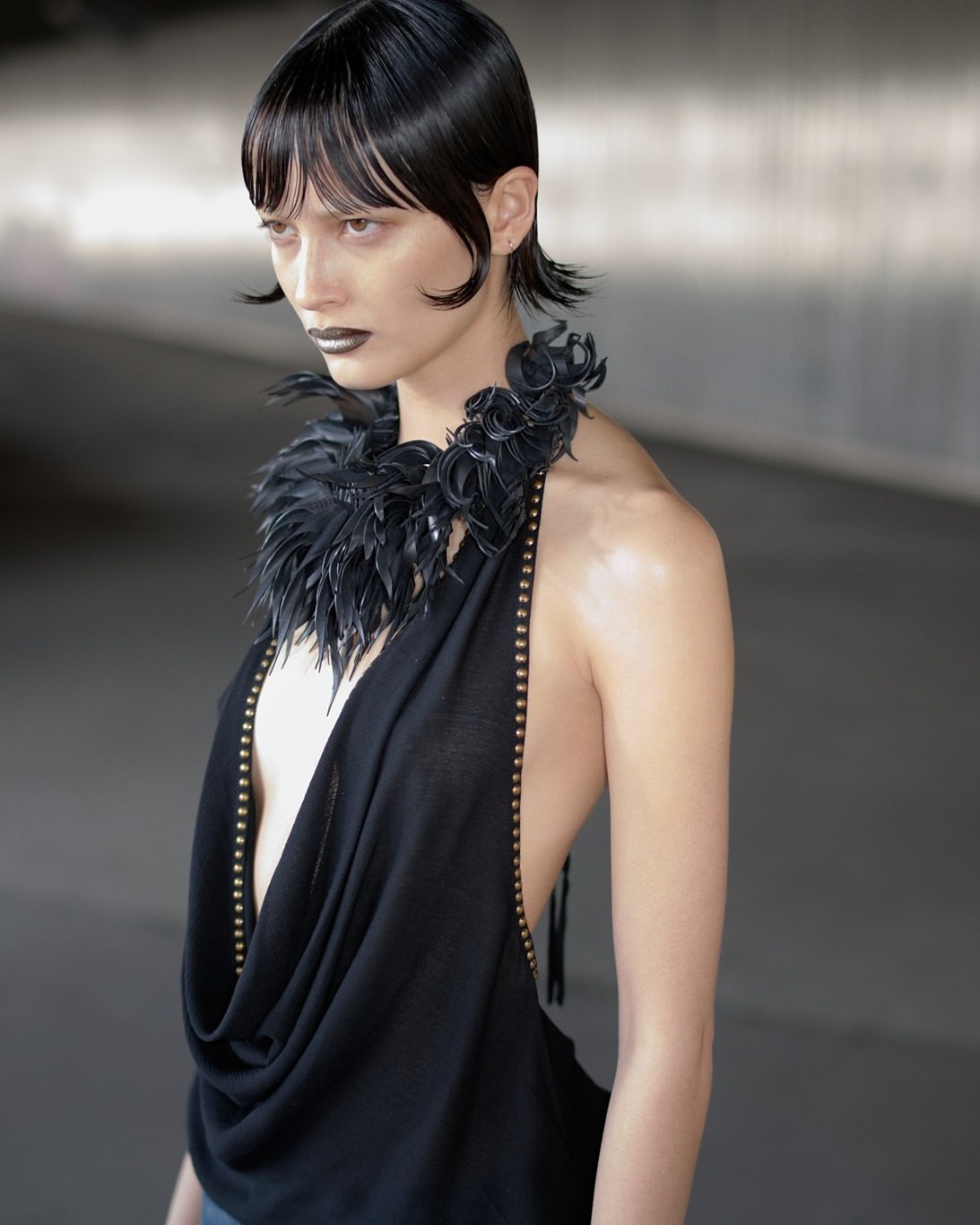 A fashionable adult woman with a sleek bob hairstyle poses wearing a black halter-top dress with a feather collar in a photo studio.
