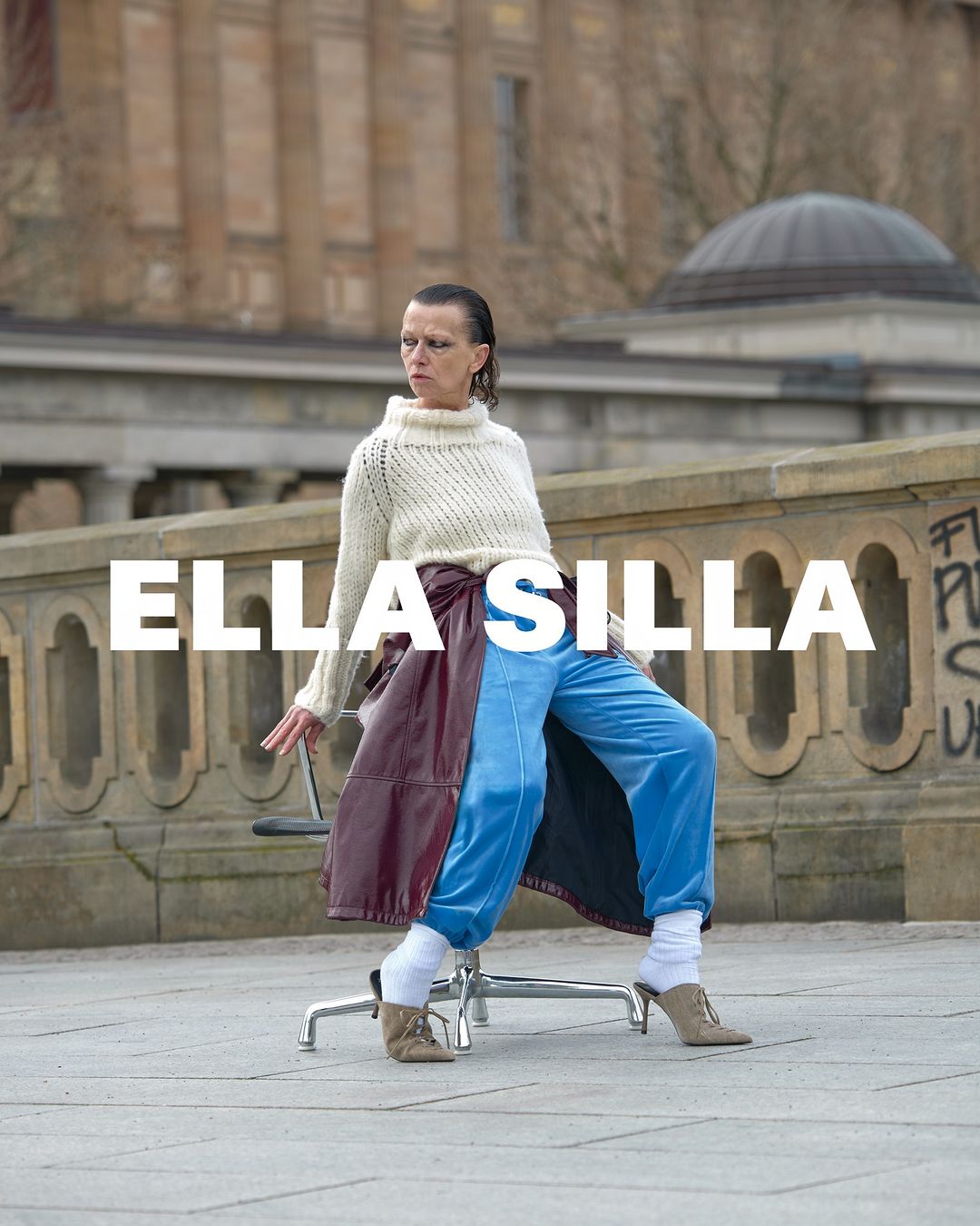 Adult female poses on a rolling chair in front of a stone building. She wears a knit sweater, blue pants, socks, and heels.