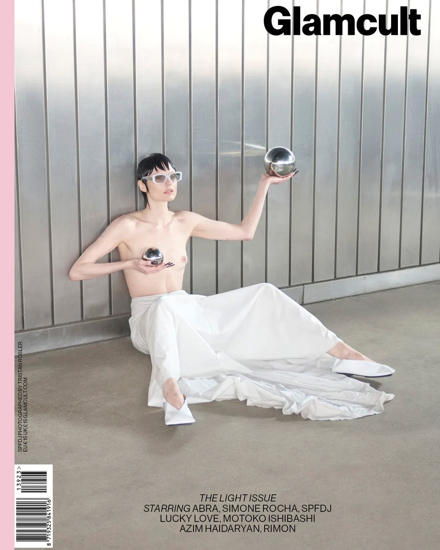 Magazine cover featuring a shirtless fashion model in white pants, sunglasses, and holding metallic spheres, posing against a metal wall.