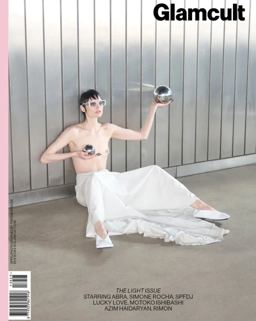 Magazine cover featuring a shirtless fashion model in white pants, sunglasses, and holding metallic spheres, posing against a metal wall.