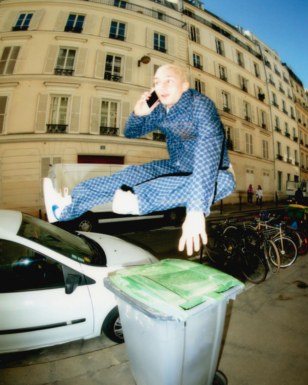 Young adult male jumping over trash can in urban setting while on phone, wearing blue patterned pajamas and white sneakers, with buildings behind.