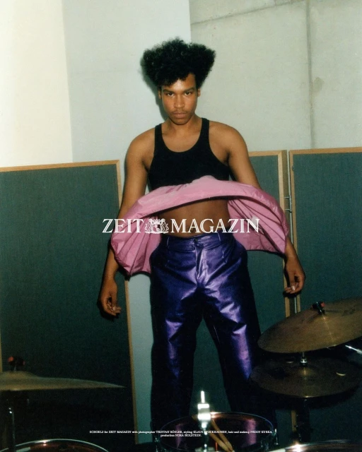 A person with an afro wearing shiny purple pants and a black tank top stands in front of a drumset and a folding privacy screen.