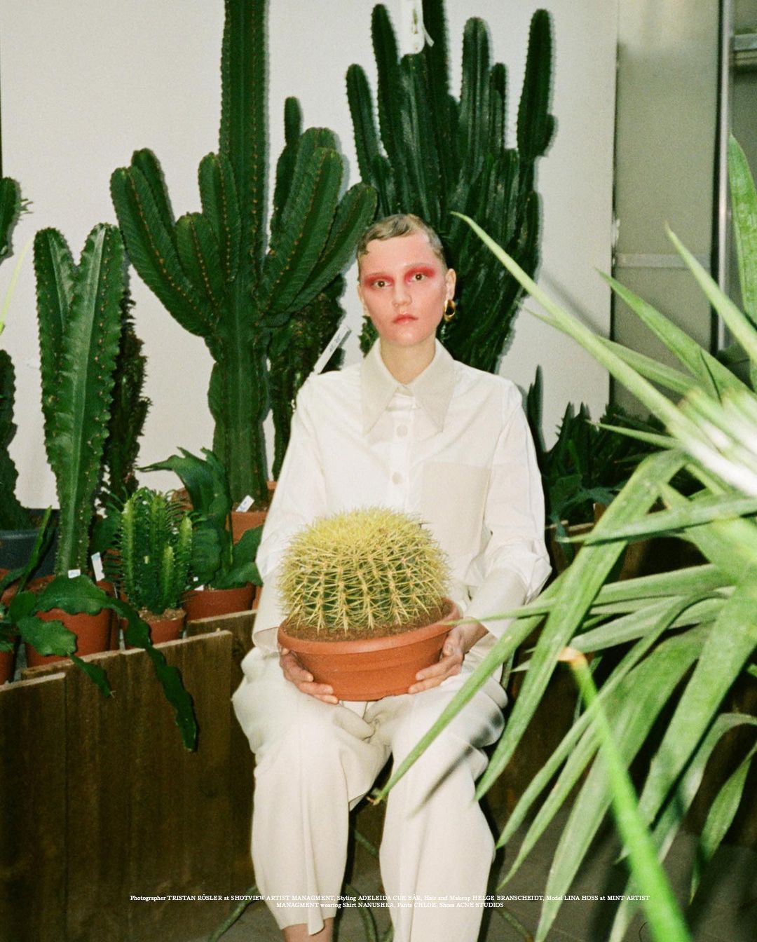 Woman in white outfit holds potted cactus in front of a green cactus display, creating a natural and styled composition.