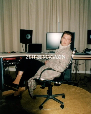 Man sits in a studio in a houndstooth coat. Studio monitors, computer, and recording equipment line the desk in the background.