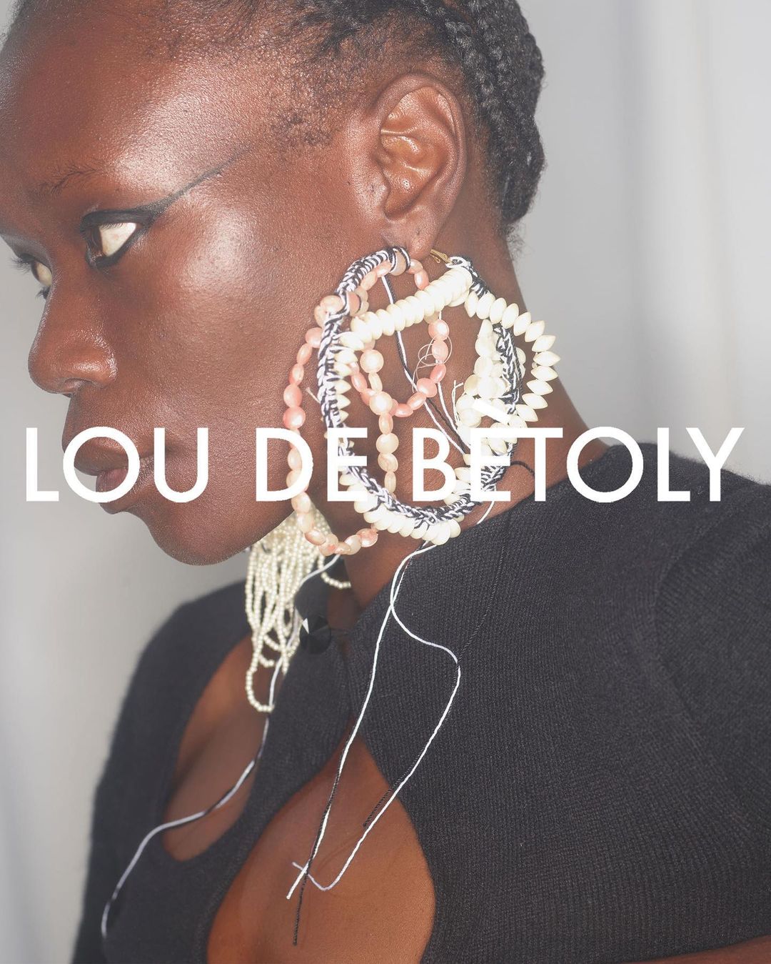 Close-up of Black female model wearing statement earrings with dramatic eye makeup and a black top against a plain background.