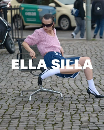 A woman in a pink sweater and blue shorts seated on an office chair outdoors on a cobblestone street in Paris photographed by Tristan Rösler.