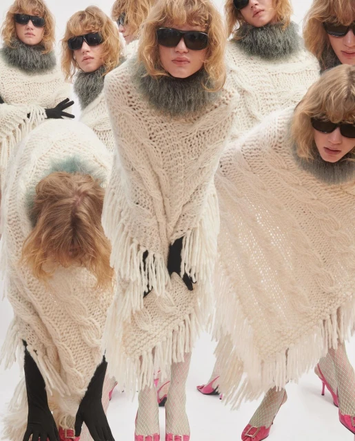Several women pose in a photo studio wearing matching cream-colored, fringe-trimmed ponchos, tights, sunglasses, and pink high heels.