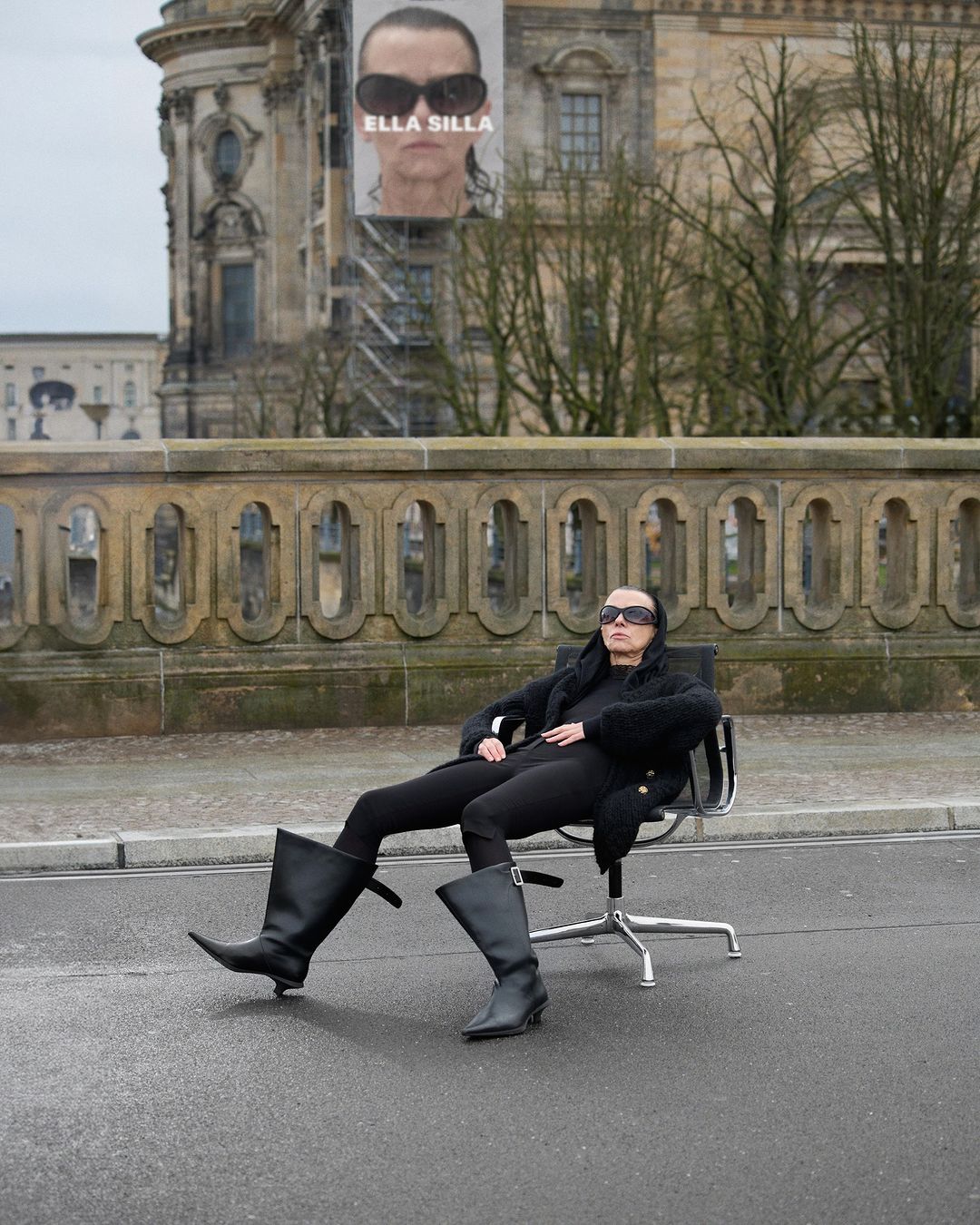 Woman in black outfit seated on an office chair on a city street in front of stone buildings. Advertising poster in background.