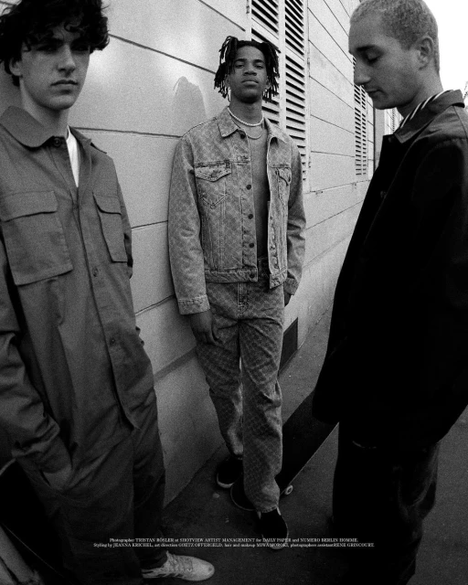 Black and white fashion shot featuring three young adult men standing against an exterior building wall wearing casual clothing.