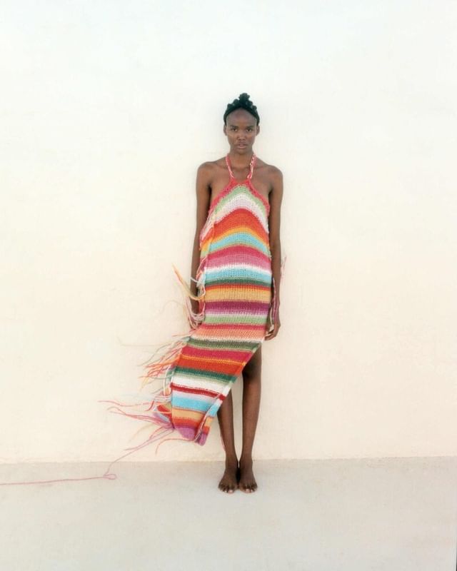 A slender Black woman stands barefoot in a photo studio, wearing a colorful, vertically striped, halter-style knit dress with fringe.