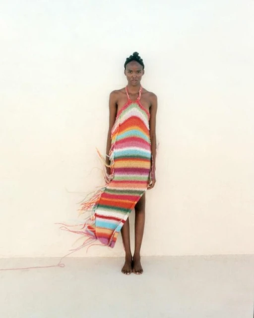A slender Black woman stands barefoot in a photo studio, wearing a colorful, vertically striped, halter-style knit dress with fringe.