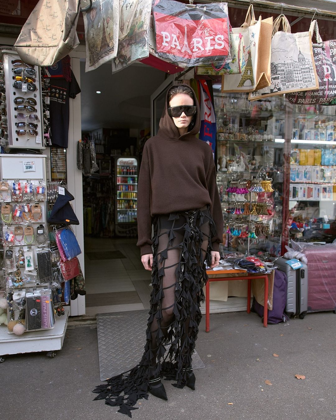 Woman with sunglasses in brown hoodie and distinctive skirt stands in front of shop with souvenirs in an urban environment on a sunny day.