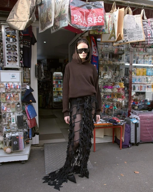 Woman with sunglasses in brown hoodie and distinctive skirt stands in front of shop with souvenirs in an urban environment on a sunny day.