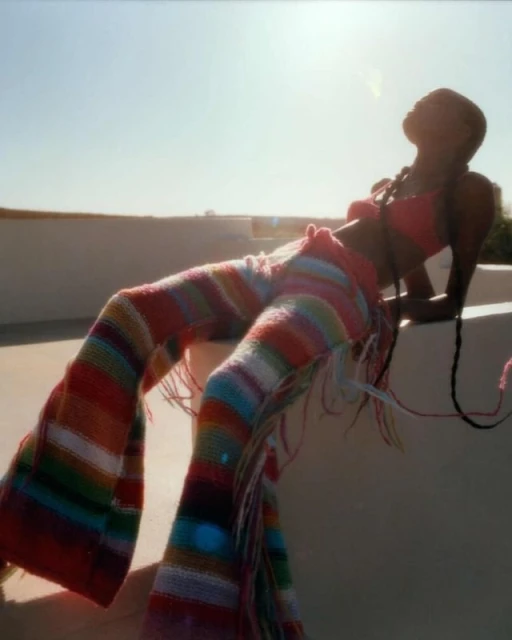 Woman in a pink top and colorful striped knit pants leans against a white wall under a sunny sky, showcasing a fashion-forward look.