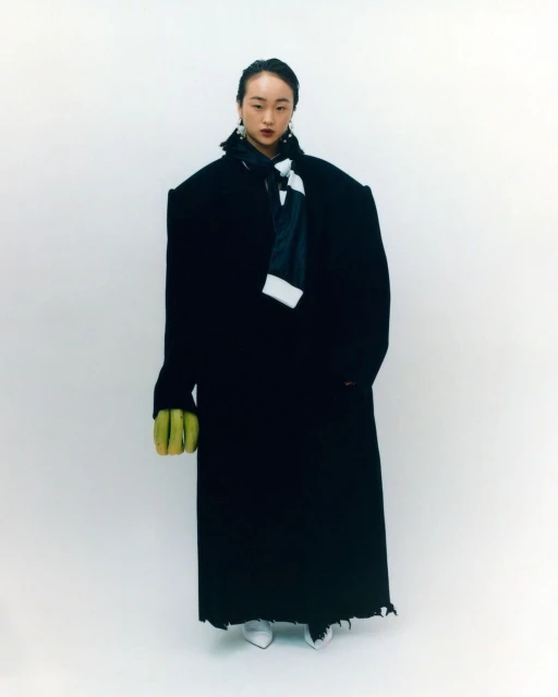 An Asian woman stands in a light studio wearing a large black coat, white shoes, and a black-and-white striped scarf, with light green gloves.