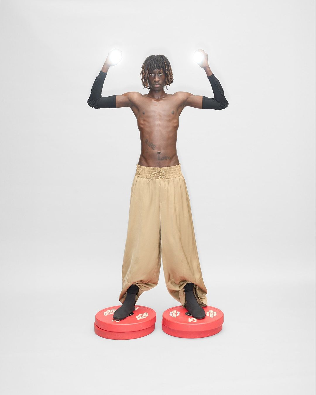 An adult male with dreadlocks poses with arms raised, lit spheres in hands, wearing wide, gold pants and standing on red circular platforms.