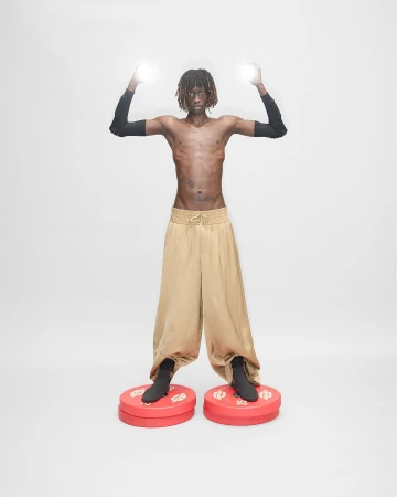 An adult male with dreadlocks poses with arms raised, lit spheres in hands, wearing wide, gold pants and standing on red circular platforms.