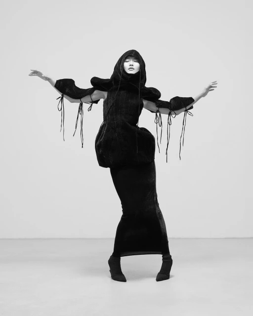 Woman in dramatic hooded couture dress strikes a pose in a stark studio. The black-and-white photograph focuses on avant-garde fashion.