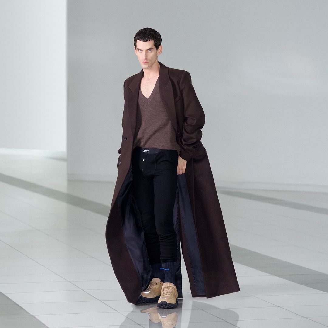 Male fashion model showcases a long brown coat, sweater, and dark pants in a minimalist studio with bright, neutral lighting.