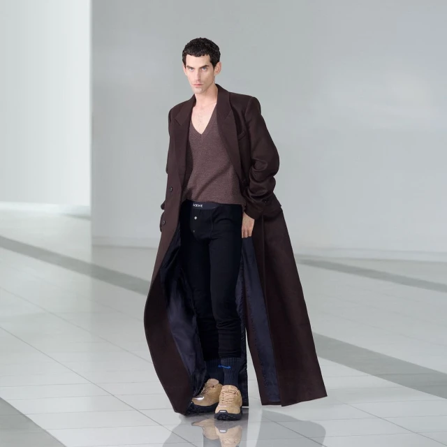 Male fashion model showcases a long brown coat, sweater, and dark pants in a minimalist studio with bright, neutral lighting.
