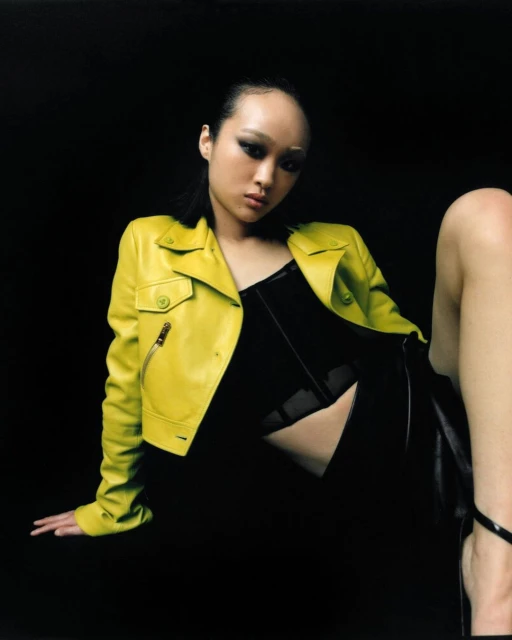 Eye-level shot of a young Asian woman in a chartreuse motorcycle jacket and black dress, seated against a dark backdrop.
